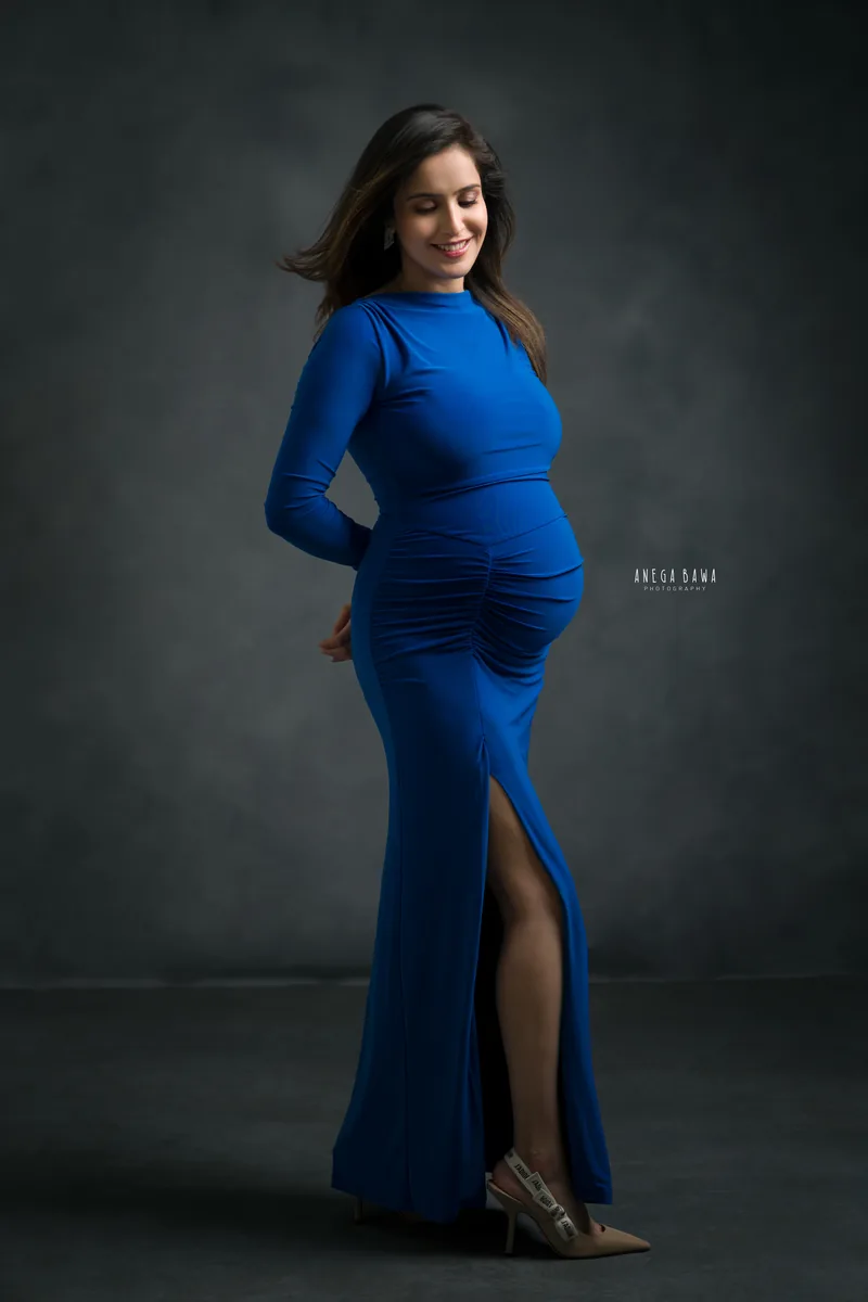 Maternity photoshoot in Delhi: expecting mother in a royal blue ruched long-sleeve gown with a side slit and nude heels, hands behind her