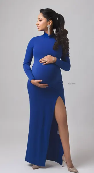 Expecting mother in a cobalt blue high-neck long-sleeve fitted gown with a thigh-high slit and nude heels, ponytail with drop earrings