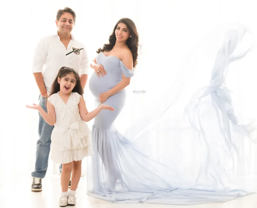 Family of three: husband in a white shirt and jeans, young daughter in a white tiered dress with arms out, wife in a pale blue off-shoulder