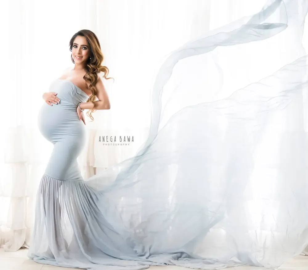 Expecting mother in a pale grey-blue strapless mermaid gown with a sheer train flying out behind her, bright white curtain backdrop, hand