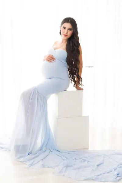 Expecting mother seated on stacked white blocks in a pale blue strapless mermaid gown with a long chiffon train, long wavy hair, hand on
