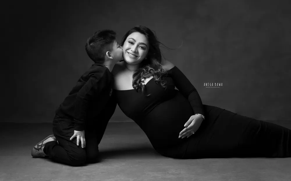 Maternity photoshoot in Delhi: black and white portrait of a young son kissing his smiling mother's cheek as she reclines in a black gown