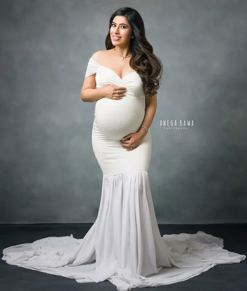 Smiling expecting mother in a white off-shoulder sweetheart mermaid gown with a wide spread train, hands on her bump, grey-blue backdrop.