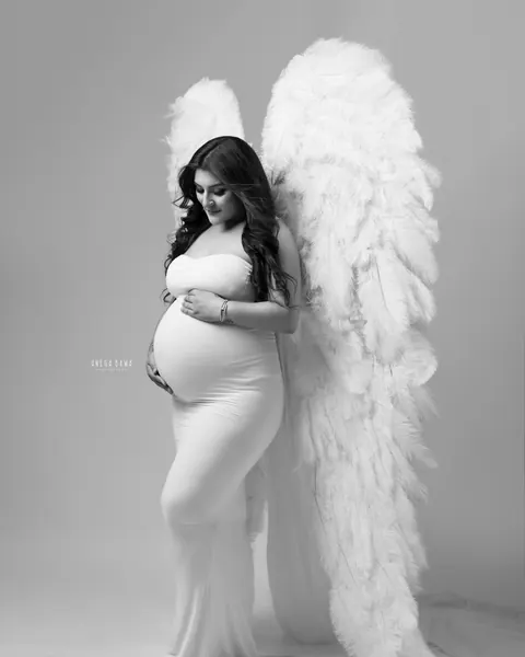 Maternity photoshoot in Delhi: black and white portrait of expecting mother in a fitted white gown standing in front of large white angel