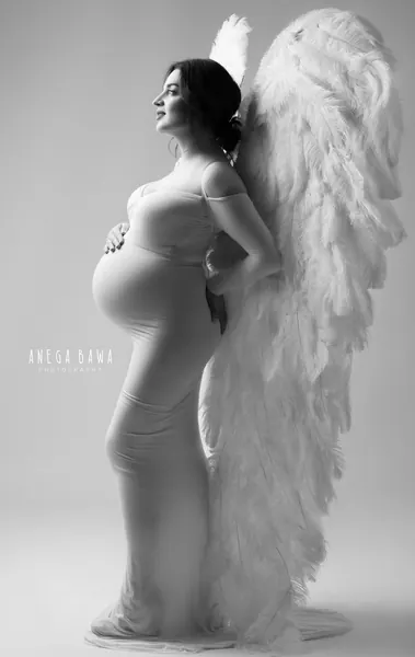 Black and white portrait of expecting mother in a fitted off-shoulder gown in profile in front of large white feather angel wings, hand on