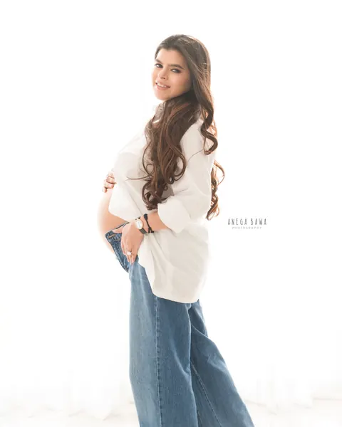 Maternity photoshoot in Delhi: expecting mother in a white shirt and wide-leg jeans standing in side profile and smiling at the camera
