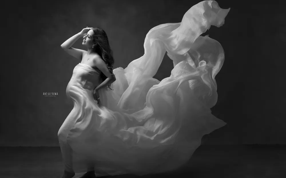 Maternity photoshoot in Delhi: black and white portrait of expecting mother in a strapless white silk drape with fabric billowing behind