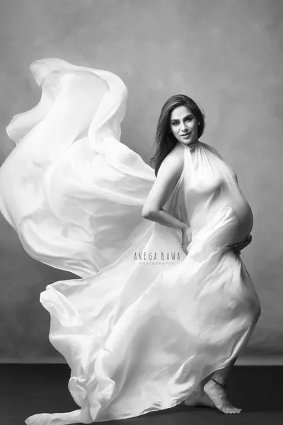 Black and white portrait of expecting mother in a white silk halter drape with fabric flying out around her, one foot lifted, hand on her