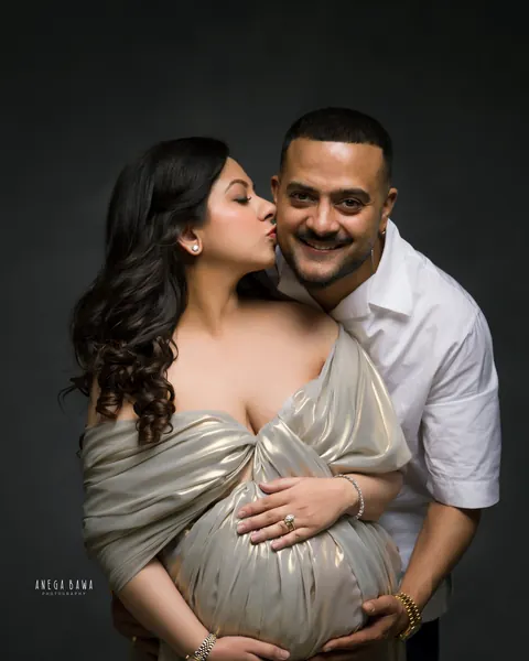 Maternity photoshoot in Delhi: expecting mother in a gold metallic drape kissing her smiling husband's cheek as he holds her bump from