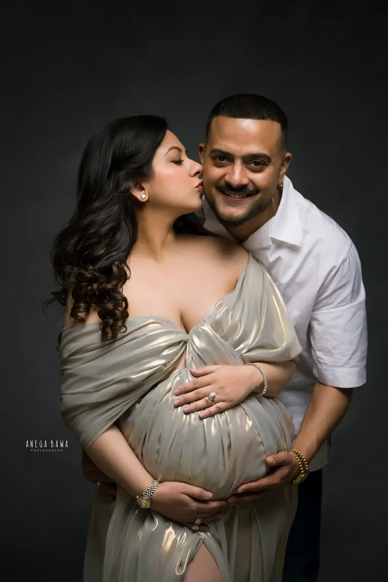 Maternity photoshoot in Delhi: expecting mother in a gold metallic drape kissing her smiling husband's cheek as he holds her bump from
