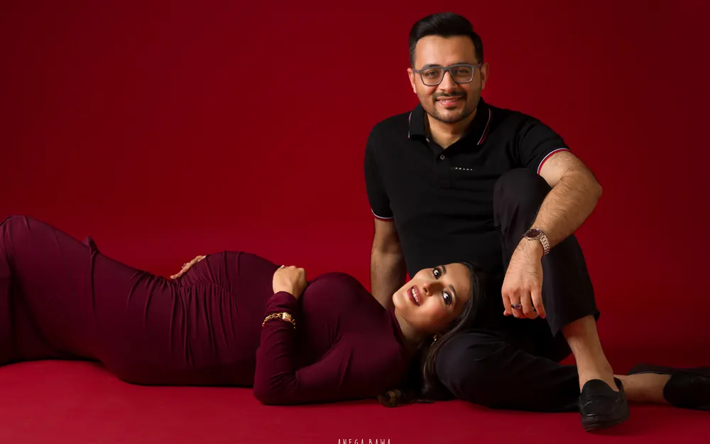 Maternity photoshoot in Delhi: expecting mother in a maroon gown lying on the studio floor with her head against her husband's leg as he