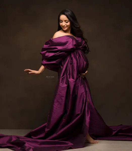 Expecting mother in a deep plum satin gown with puffed off-shoulder sleeves wrapped around her bump and a long train on the floor