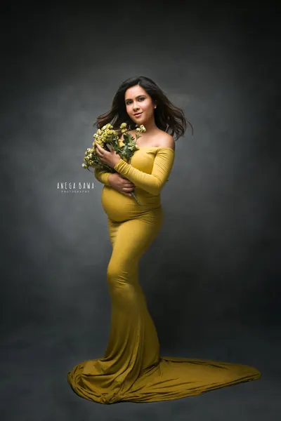 Expecting mother in a mustard yellow off-shoulder long-sleeve mermaid gown with a train, holding a bunch of yellow flowers, dark moody