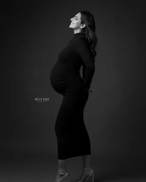 Maternity photoshoot in Gurgaon: black and white profile of expecting mother in a black high-neck dress and heels, face lifted with a smile