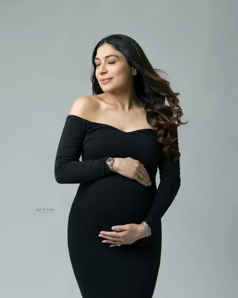 Maternity photoshoot in Gurgaon: expecting mother in a fitted black off-shoulder gown with eyes closed and both hands on her bump, soft