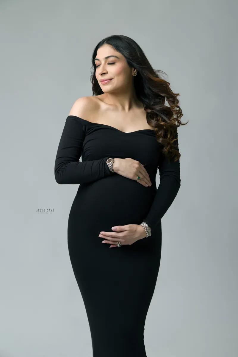 Maternity photoshoot in Gurgaon: expecting mother in a fitted black off-shoulder gown with eyes closed and both hands on her bump, soft