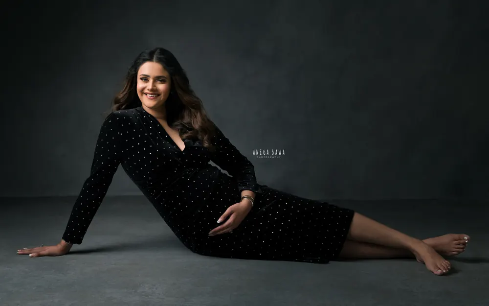 Maternity photoshoot in Gurgaon: smiling expecting mother reclining barefoot on the studio floor in a black velvet wrap dress with silver
