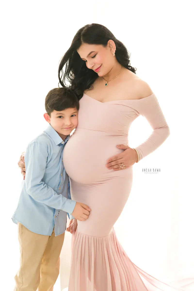 Maternity photoshoot in Gurgaon: expecting mother in a blush pink off-shoulder mermaid gown with her young son leaning against her bump