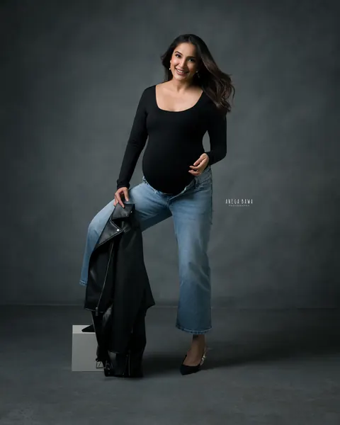 Maternity photoshoot in Gurgaon: smiling expecting mother in a black bodysuit and cropped jeans, one foot on a block and a leather jacket