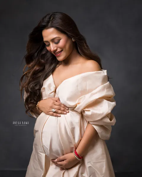 Maternity photoshoot in Gurgaon: smiling expecting mother in a champagne taffeta off-shoulder gown, hands on her bump, dark backdrop, by