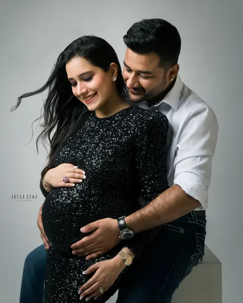 Maternity photoshoot in Gurgaon: smiling couple, wife in a black sequin gown and husband in a white shirt hugging her bump from behind