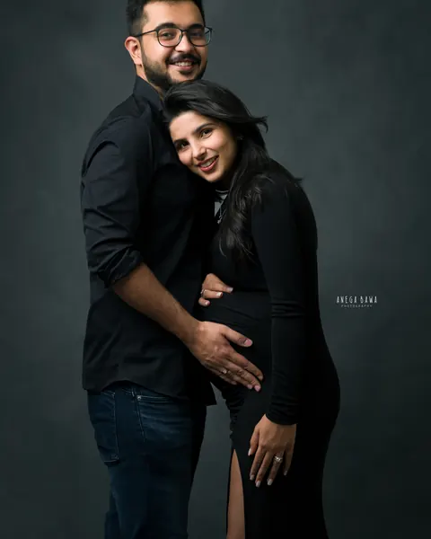 Maternity photoshoot in Gurgaon: smiling couple in black outfits, husband's hand on her bump, she wears a black gown with a side slit, dark