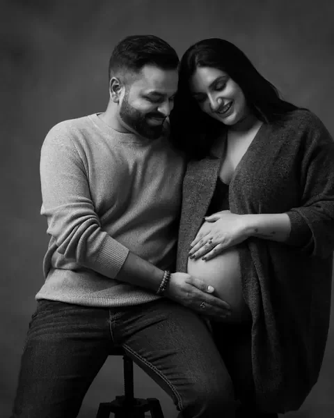 Maternity photoshoot in Gurgaon: black and white portrait of a couple seated on a stool looking down at the bump, wife in an open cardigan