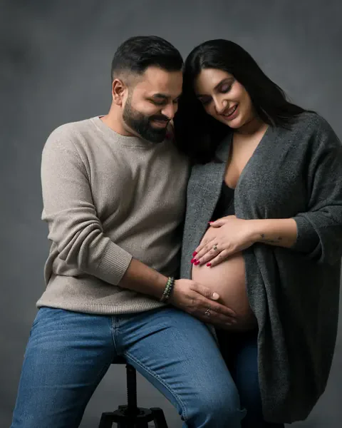 Maternity photoshoot in Gurgaon: couple seated on a stool looking down at the bump, husband in a beige sweater and wife in an open grey