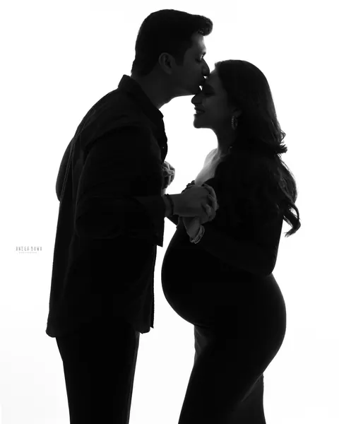 Maternity photoshoot in Gurgaon: black and white silhouette of a husband kissing his wife's forehead as they hold hands, her bump in