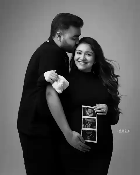 Maternity photoshoot in Gurgaon: black and white portrait of a husband kissing his smiling wife's forehead while she holds ultrasound scan