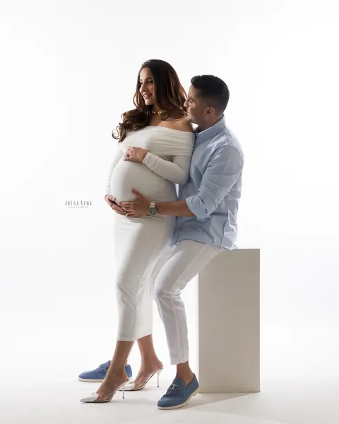 Maternity photoshoot in Gurgaon: husband in a light blue shirt seated on a white block hugging his wife in a fitted white off-shoulder
