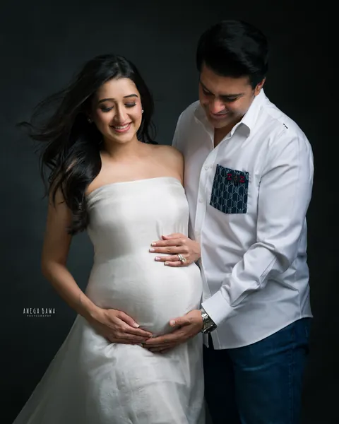 Maternity photoshoot in Gurgaon: expecting mother in a strapless white satin gown with her husband in a white shirt, both looking down at