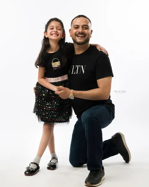 Maternity photoshoot in Gurgaon: laughing father kneeling beside his young daughter, both in black, white studio backdrop, by Anega Bawa