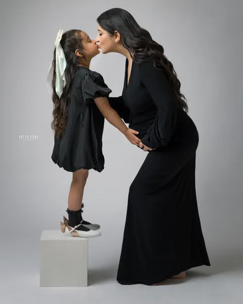 Maternity photoshoot in Gurgaon: young daughter in a black puff-sleeve dress and large bow standing on a white block to kiss her expecting