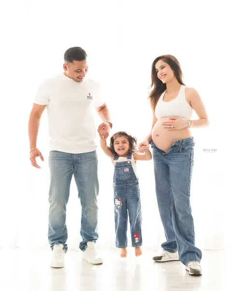 Maternity photoshoot in Gurgaon: parents in white tops and jeans swinging their laughing toddler between them, mother's bare bump showing