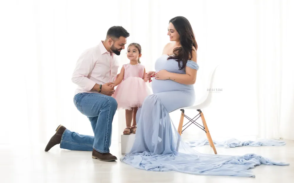 Maternity photoshoot in Gurgaon: expecting mother seated on a white chair in a powder blue off-shoulder gown with a long sheer train
