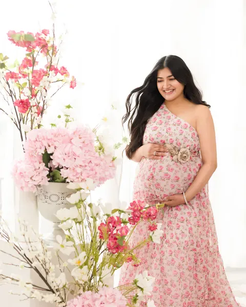 Maternity photoshoot in Gurgaon: smiling expecting mother in a pink floral-print one-shoulder dress with a flower belt, surrounded by