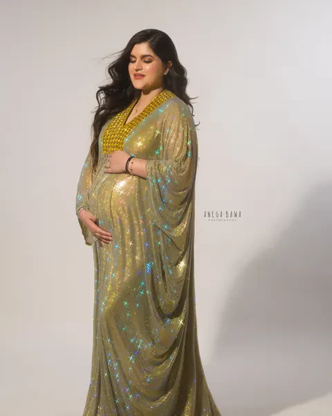 Maternity photoshoot in Gurgaon: full-length portrait of expecting mother with eyes closed in a sparkling gold kaftan gown with draped