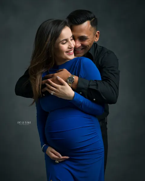 Maternity photoshoot in Gurgaon: husband hugging his laughing wife from behind, she wears a royal blue gown and cradles her bump, dark