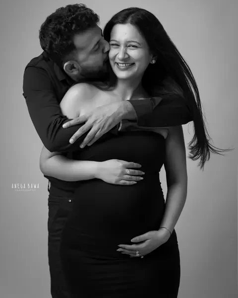 Maternity photoshoot in Gurgaon: black and white portrait of a husband kissing his laughing wife's cheek from behind, both in black, hands