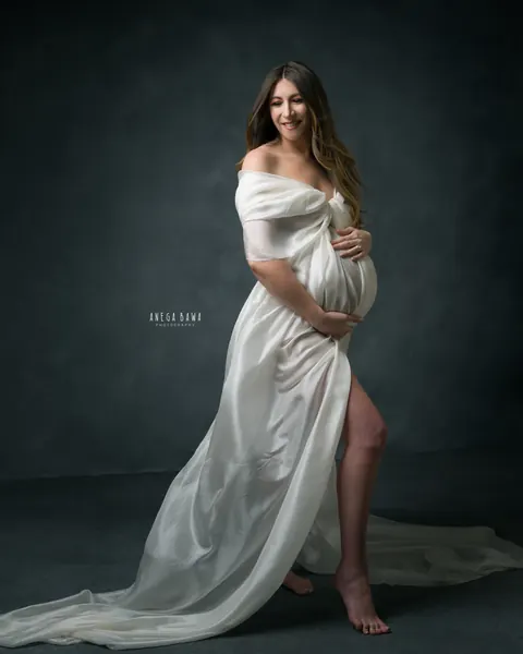 Maternity photoshoot in Gurgaon: expecting mother in a flowing ivory silk drape with a long train and side slit, slate blue studio