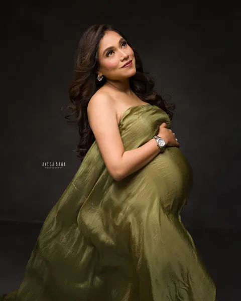 Maternity photoshoot in Gurgaon: expecting mother in an olive green satin drape gazing upward with her hand on her bump, dark backdrop, by