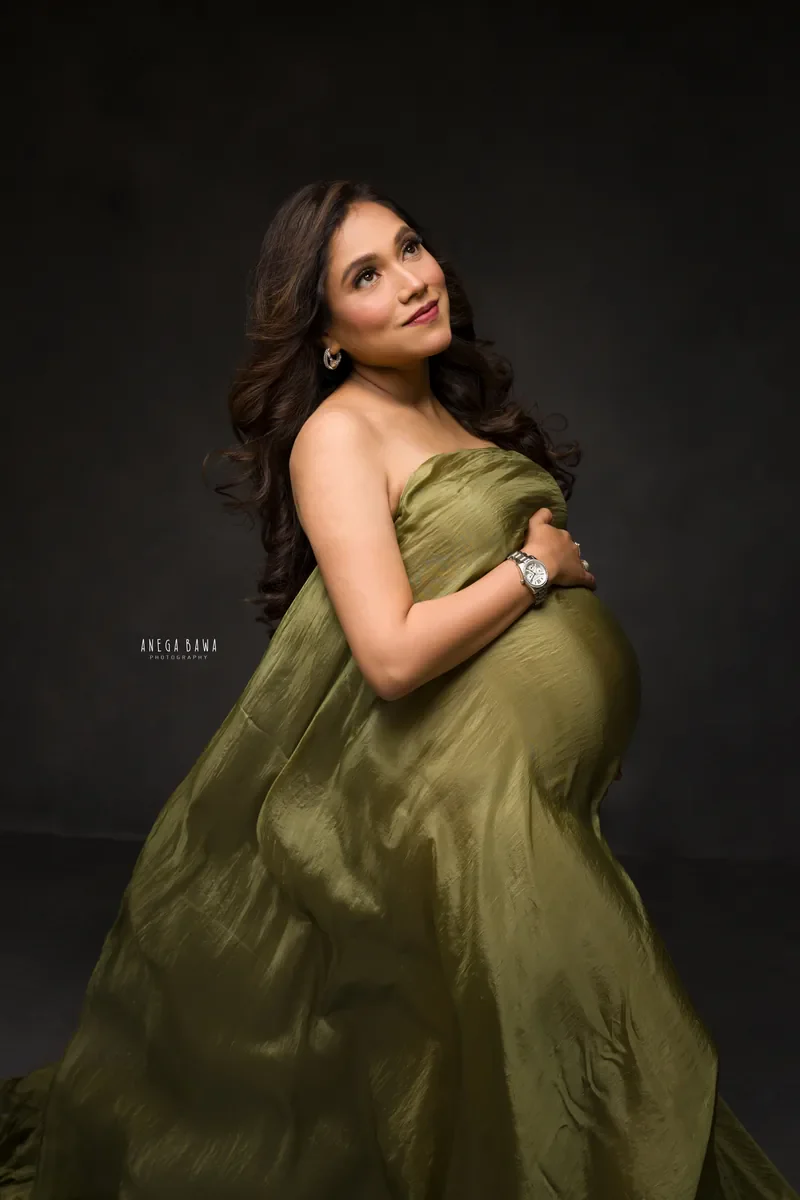 Maternity photoshoot in Gurgaon: expecting mother in an olive green satin drape gazing upward with her hand on her bump, dark backdrop, by