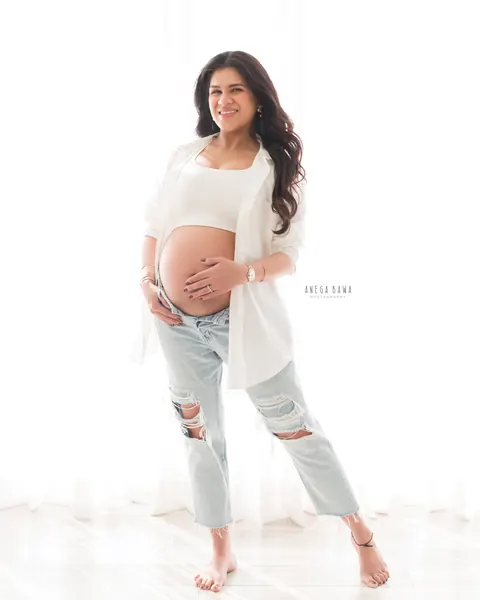 Maternity photoshoot in Gurgaon: smiling expecting mother standing barefoot in an open white shirt and distressed light denim with a hand
