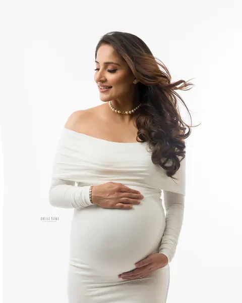 Maternity photoshoot in Gurgaon: smiling expecting mother in a fitted white off-shoulder gown and pearl necklace, hair blowing, hands on