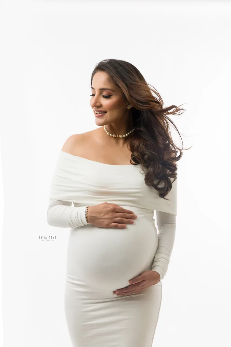 Maternity photoshoot in Gurgaon: smiling expecting mother in a fitted white off-shoulder gown and pearl necklace, hair blowing, hands on