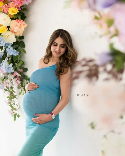 Maternity photoshoot in Gurgaon: expecting mother in a fitted teal one-shoulder gown leaning on a white wall dressed with pastel flowers