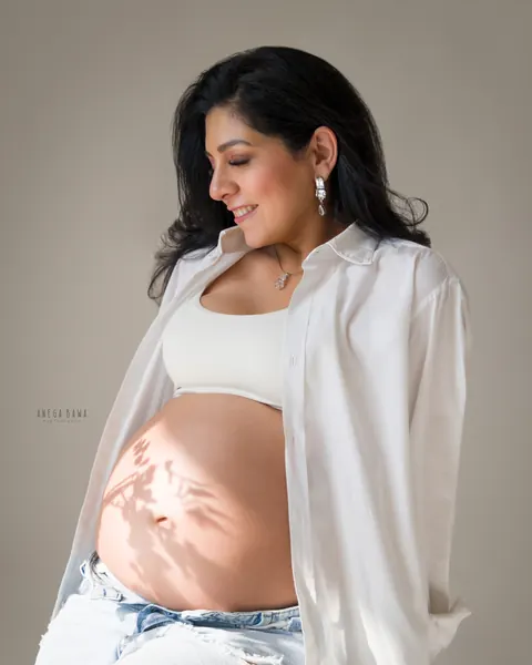 Maternity photoshoot in Gurgaon: expecting mother in an open white shirt and light denim with soft window light and leaf shadows on her