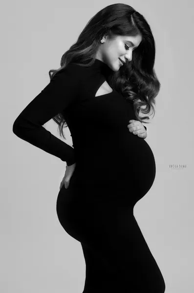 Black and white portrait of expecting mother in profile in a black long-sleeve cut-out dress, looking down at her bump, hand on her lower