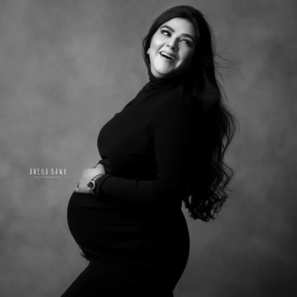 Black and white portrait of laughing expecting mother in a black turtleneck fitted outfit with long wavy hair, looking back over her